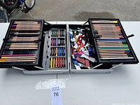 Metal art case open with colored pencils, oil pastels, and tubed paints spread in three tiers.