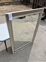 Full view of large rectangular framed mirror standing upright outdoors on pavement next to a white table.