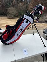 Photo of youth golf clubs in a red, white, and black Precise branded stand bag propped on a golf table outside.