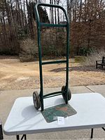 Full view of green metal hand truck standing on a white table outdoors, showing upright handle and base plate.