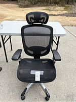 Full front view of the black mesh ergonomic computer chair placed outdoors on pavement with a folding table behind it.