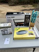 Photo showing the full lot on a table outdoors, includes Chefman grill, crock-pot in box, Emeril cast iron sizzle plate, knife block with 6 turquoise knives, and barware cocktail shaker set.