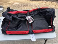 Large black and red rolling duffle bag laid on table, showing front pockets and handles, with a tag attached.