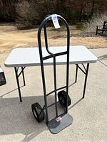 Full view of Milwaukee metal hand truck standing upright against a folding table outdoors on pavement.