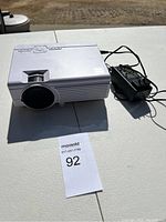 White portable projector shown from front and side angles, placed on white folding table outdoors, includes visible lens and control buttons.