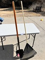 Full length view of a shovel with a brown wooden handle and a black metal spade, and a leaf rake with a cream wooden handle and black metal tines, both standing upright on a white table outdoors.