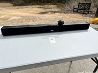 Long black RCA soundbar on white table outdoors with power cord grouped on top.