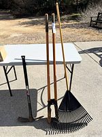 Full view of three yard tools laid on a white folding table outdoors including a post hole digger, a pick axe with a grip handle, and a plastic leaf rake with a wooden handle.