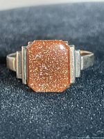 Front close-up of the rectangular faceted goldstone set in the ring, showing glittering reddish-brown color and gold and silver band details.