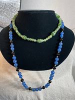 Both necklaces shown on black necklace display stand, showing full length and color contrast of blue black necklace and green glass choker.