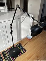 Full view of black Ikea floor standing reading lamp by kitchen counter on wooden floor showing weighted base, adjustable arm, and lamp head.