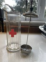 Clear view of vintage glass Barbicide jar with red cross and metal plunger lid removed, showing perforated basket inside.