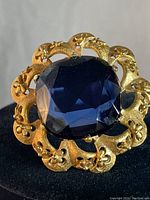 Front view showing the deep blue cushion-cut faceted glass stone set in ornate gold plated floral frame.