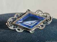 Close-up front view of ornate sterling silver brooch with faceted elongated diamond-shaped blue stone.