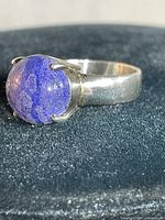 Close-up of sterling silver ring with round polished blue Larus Lazulli stone set in prongs on a black fabric background.