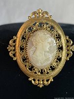 Front view of vintage Victorian gold tone ornate cameo brooch with intricate filigree frame and profile cameo of woman.