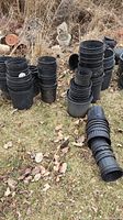 Multiple stacks of black plastic potting containers in various sizes outside on grass with dry vegetation background.