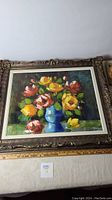 Full view of framed oil painting featuring colorful flowers in a blue vase on a dark background with ornate frame.