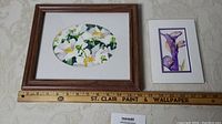 Two framed watercolor paintings, one larger floral oval by M. Rutledge with white flowers and the other a smaller purple iris painting, pictured with ruler for scale.