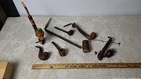 Overview of 7 vintage old pipes arranged on table with ruler for scale, showing different shapes and materials