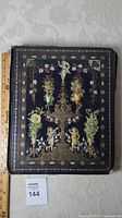 Closed view of vintage scrapbook with decorative black cover featuring cherubs and floral motifs.