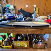 Photo showing assortment of cleaning fluids, wax, microfiber towels, sponges, and automotive maintenance products organized in boxes under and on a wooden table. Floor mats and trunk covers also visible behind.