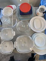 Top view of lot showing multiple Corning Ware casseroles with lids, Pyrex glassware, Corelle plates and bowls, and a pitcher with floral decoration.