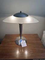 Front view of modern oval table lamp on wooden surface, showing brushed steel base and frosted glass shade with top steel dome.