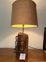 Front view of tiki style vintage driftwood lamp lit, showing carved face and metal chain on base, with woven brown drum shade.