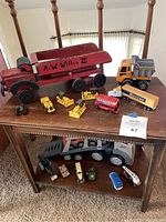 Photo of the full lot showing a large vintage wooden truck, multiple small yellow construction vehicles, an orange dump truck, a Baxter semi trailer, a red double-decker bus, various small toy vehicles including cars and a helicopter, and a large toy truck with trailer on the lower shelf.