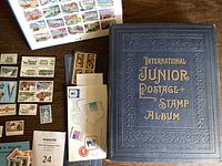 Photo of a blue hardcover International Junior Postage Stamp Album, unopened, alongside various individual stamps and stamped envelopes laid out on a table.