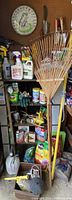 Photo of a wooden rake with a yellow handle, metal hand garden tools hanging on a wall, assorted bottles of garden chemicals and sprays on shelves.