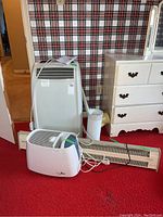 Photo showing the Noma portable air conditioner, Sentinel floor heater, and Germ Free humidifier arranged on red carpet with wallpaper background. Manual placed on air conditioner.