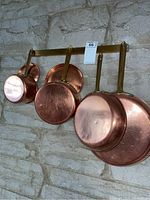 Overall view of two fry pans and three lidded pots hanging on brass rack against stone wall