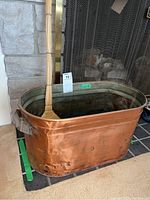 Front side view of the large rectangular copper tub with a small broom inside it, showing overall wear and one broken handle.