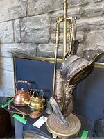 Full view of the fireplace stand with four brass tools, two miniature teapots, and a bed pan on a dark surface against stone wall.