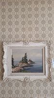Front view of framed island scene painting hung on patterned wallpaper showing ornate white carved wood frame and seascape with rocky island and trees.