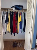 Closet view showing a variety of men’s button-down shirts, polo shirts, and jackets hanging on a rod with several pairs of folded pants on the shelf above.