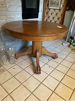 Round solid oak dining table with pedestal base and claw feet on casters, wood grain visible with some surface stains.