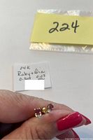 Photo showing a hand holding the pair of 14 kt gold ruby and diamond stud earrings with a label indicating stone carat weights.