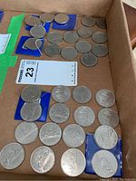 Coins shown inside a cardboard box, some coins loose and others in small blue coin holders. Coins are silver-colored one dollar pieces with visible Voyageur design.