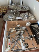 Photo of assortment of silver serving ware including flatware pieces and lidded dishes in a box and on display surfaces