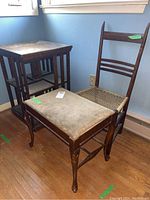 Photo showing a wooden table with square top and a wood chair with woven seat and a vintage cushioned stool in between them, placed on a hardwood floor near a blue wall.