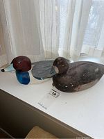 Two wood duck decoys side by side on a white surface near a window, one visibly brighter and more detailed, the other darker and worn.
