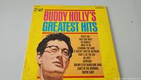 Front cover of Buddy Holly's Greatest Hits vinyl record album, yellow background with Buddy Holly's portrait and song titles in colorful text.