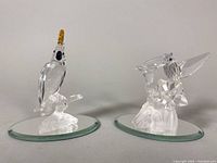 Two Swarovski crystal bird figurines on circular beveled mirrored glass bases: parrot and hummingbird.