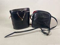 Two black leather cases, one a larger square with top opening and strap, the other a smaller, rounded pouch style with red fabric interior visible, for binoculars.
