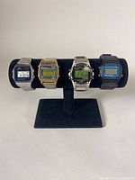 Front view of four men's digital watches on black display stand showing faces and metal/plastic bands.