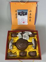 Photo of tea set box open showing teapot, cups, and saucers wrapped partially in paper in yellow satin lining with certificate and calligraphy inside lid.