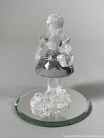 Front view of the crystal little girl figurine standing on a round beveled mirrored base, showing the detailed face and basket.